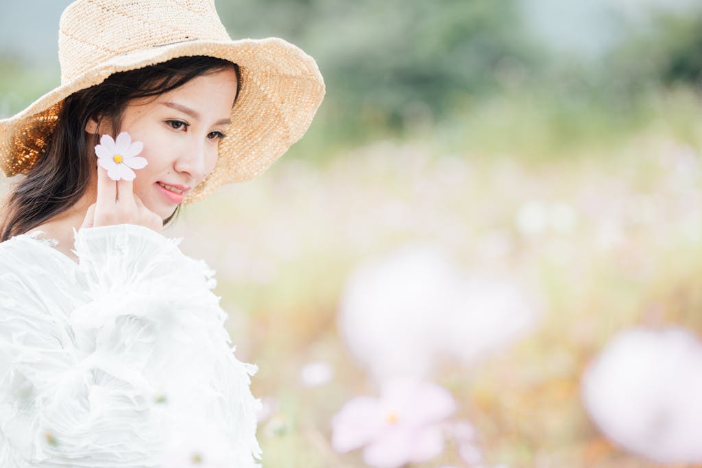 A woman in a straw hat and white dress holds a flower, enjoying a sunny day in a bright, idyllic field.