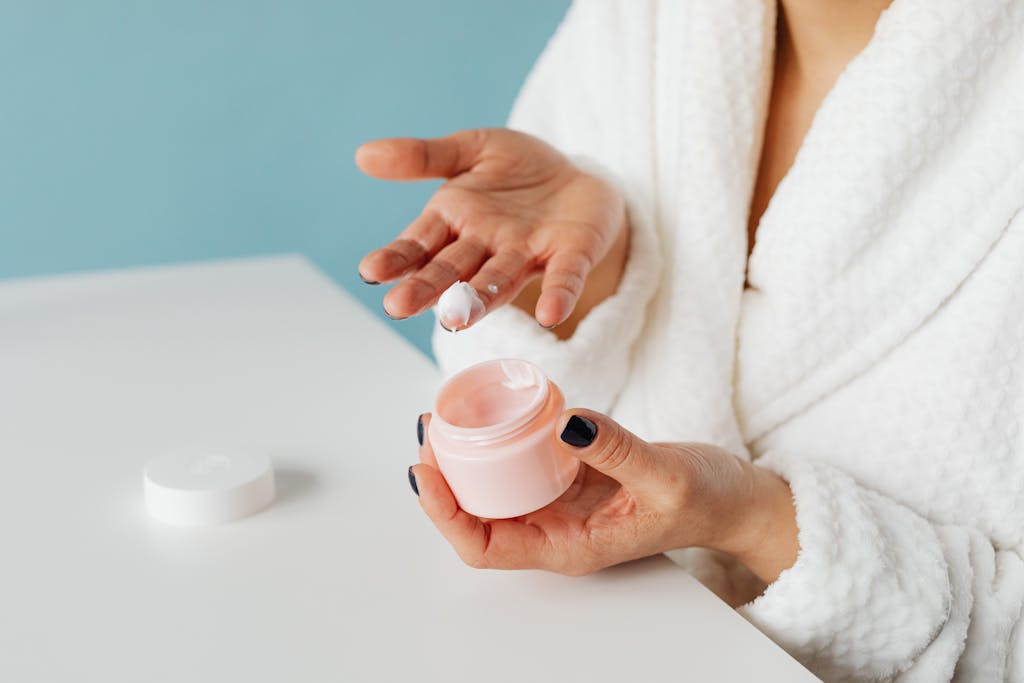 A woman in robe applying skincare cream from an open jar, highlighting moisturizing routine.