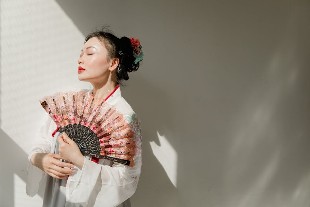 A woman in traditional hanfu poses elegantly with a decorative hand fan in soft natural light.