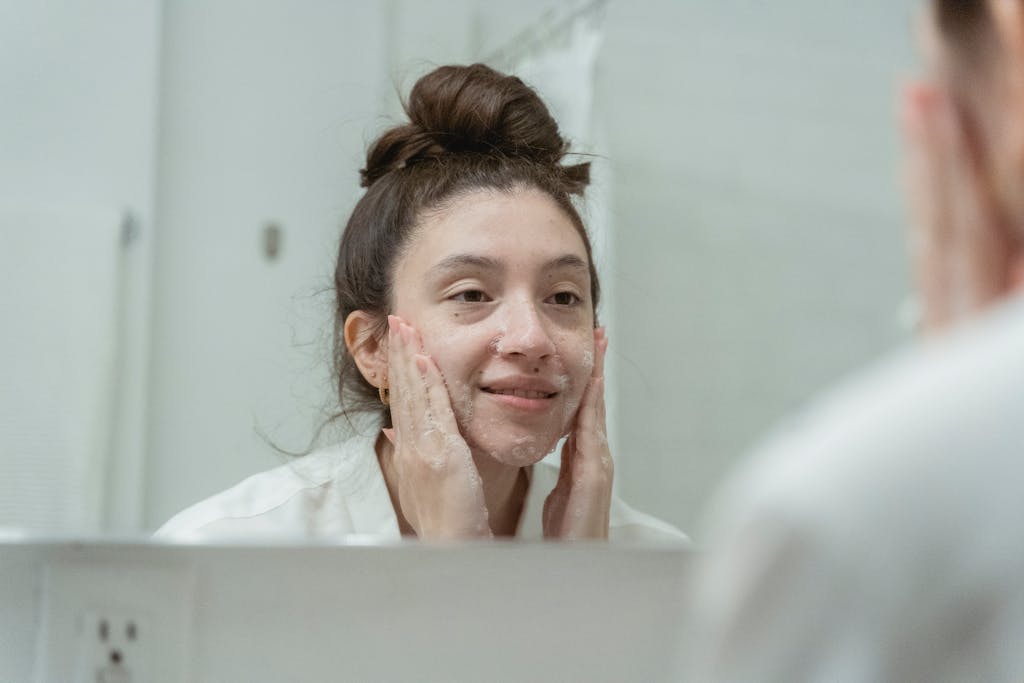 A woman smiling as she applies facial cream in front of a bathroom mirror, enhancing skincare routine.