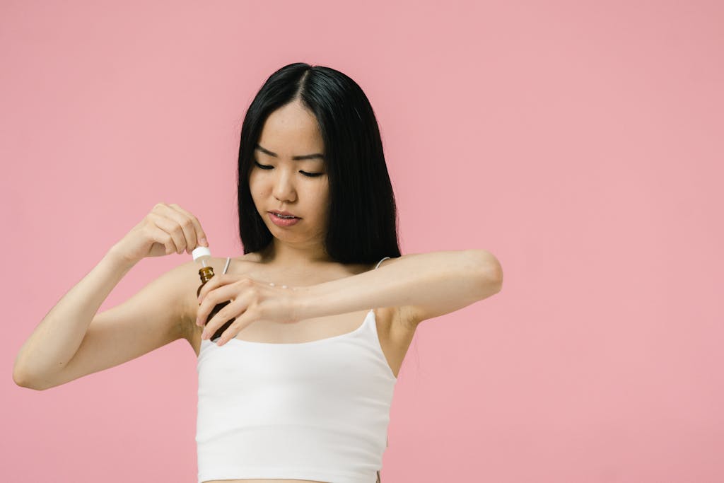 Asian woman using skincare product with pipette against a pink background, promoting self-care and wellness.