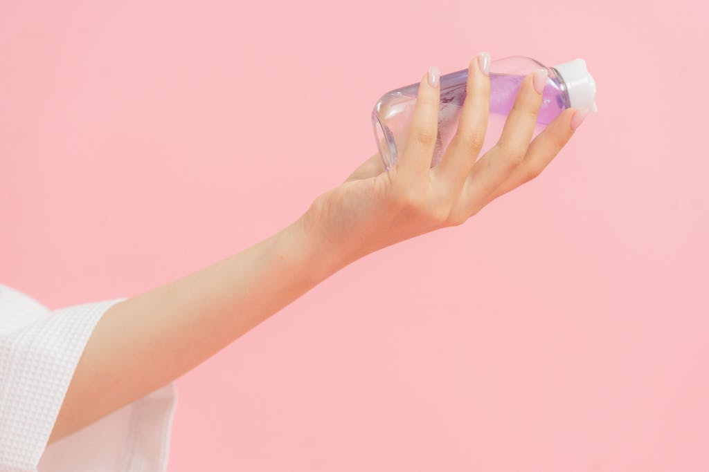 Close-up of a hand holding a clear plastic skincare bottle against a pastel pink background.