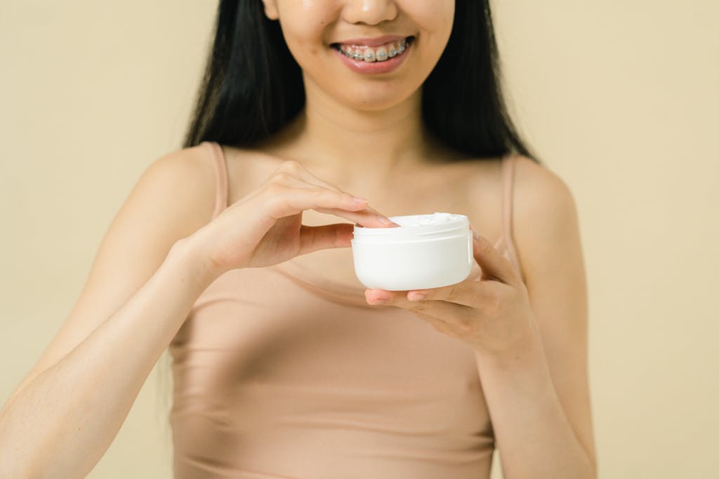 Close-up of a smiling woman applying cream from a container, showing smooth skin and braces.