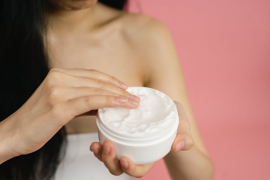 Close-up of a woman applying white cream from a jar against a pink background.