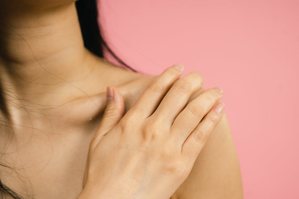 Close-up of a woman's hand resting on her shoulder against a pink backdrop, highlighting skin and nails.