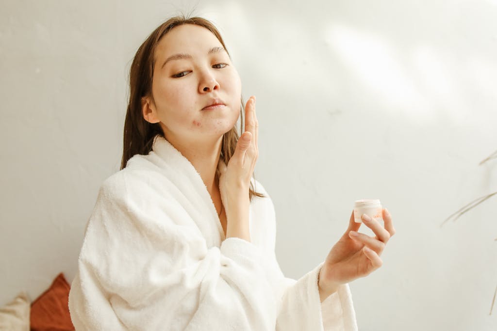 Close-up of an Asian woman in a bathrobe applying skincare cream as part of her beauty routine.