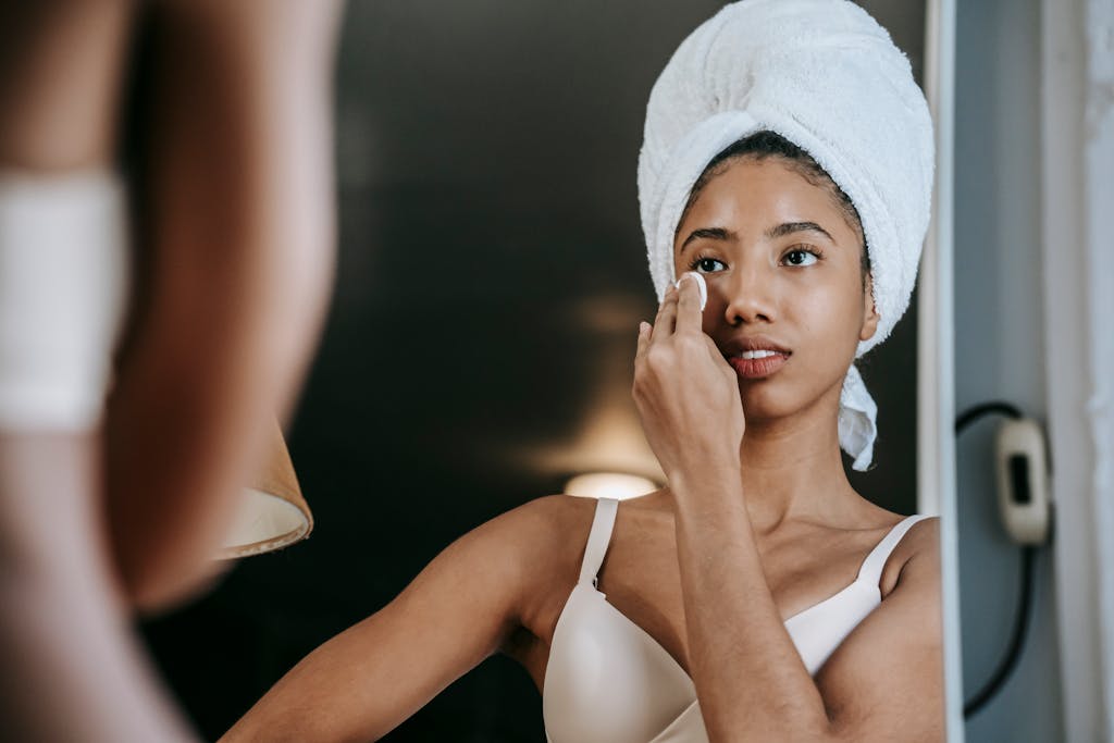 Crop young ethnic female in terry towel and brassiere cleaning face with cotton sponge while looking in mirror at home