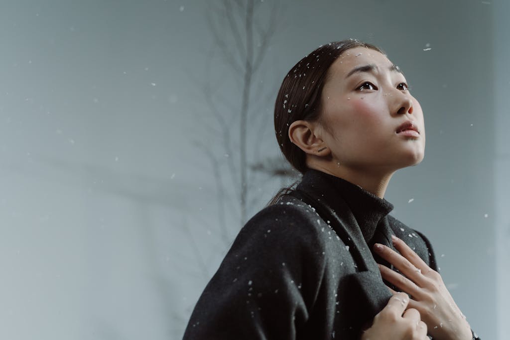Elegant Asian woman wearing a black coat looks upwards as snowflakes gently fall around her.
