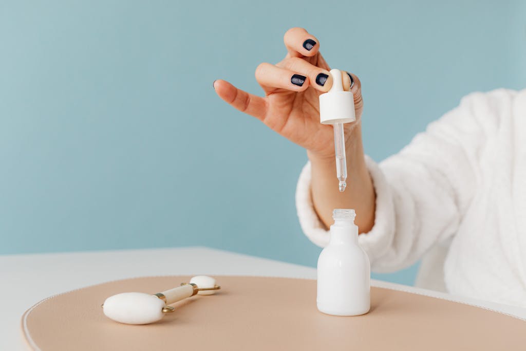 Hand in a bathrobe using a pipette to dispense skincare product into a white bottle.