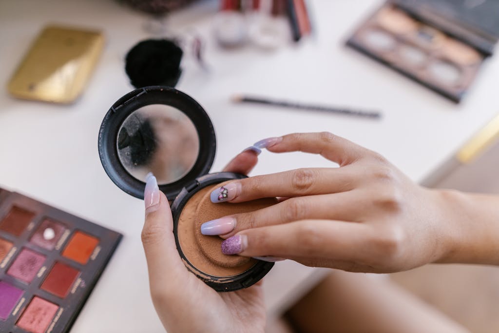 Hands applying powder with a sponge from a compact, surrounded by makeup products.