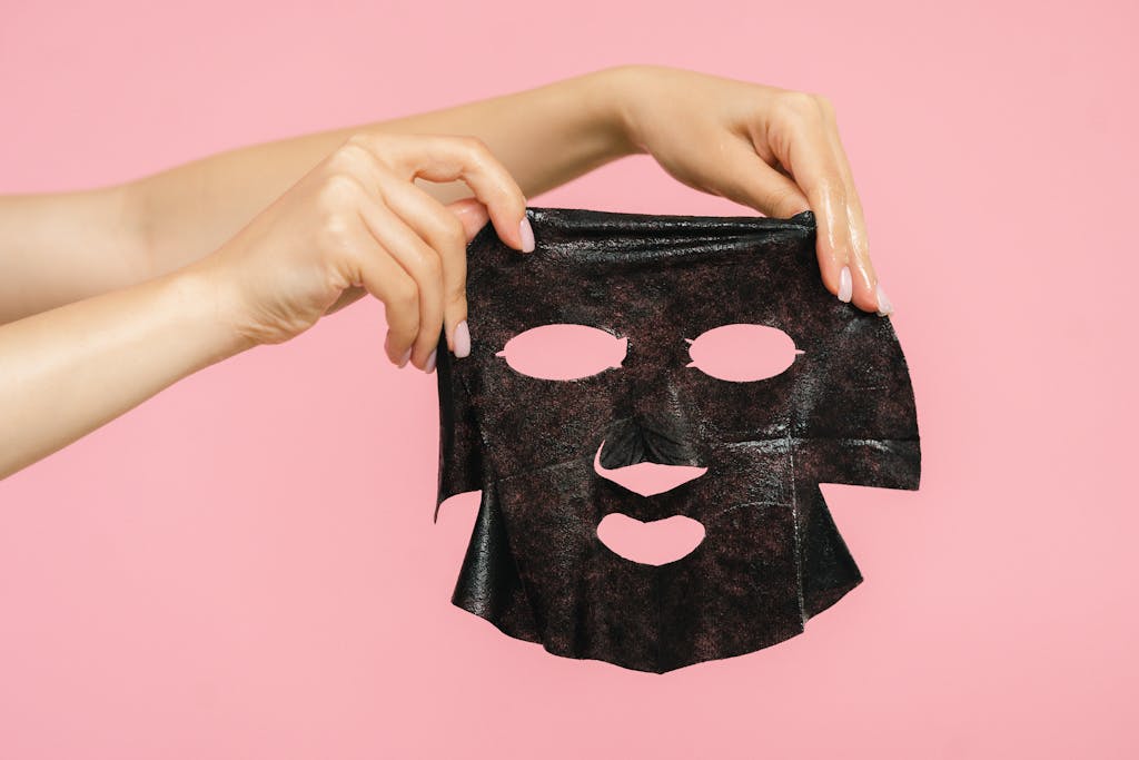 Hands hold a black charcoal facial mask against a pink background, emphasizing skincare.