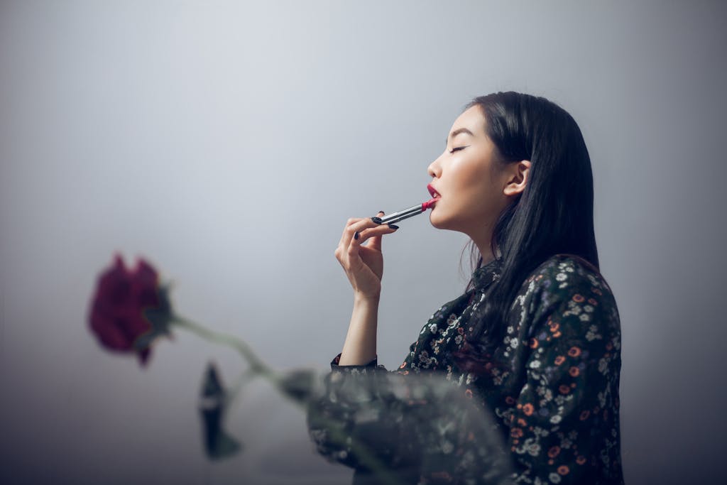 Side view of Asian woman in trendy clothes putting lipstick on lips while standing near rose in vase