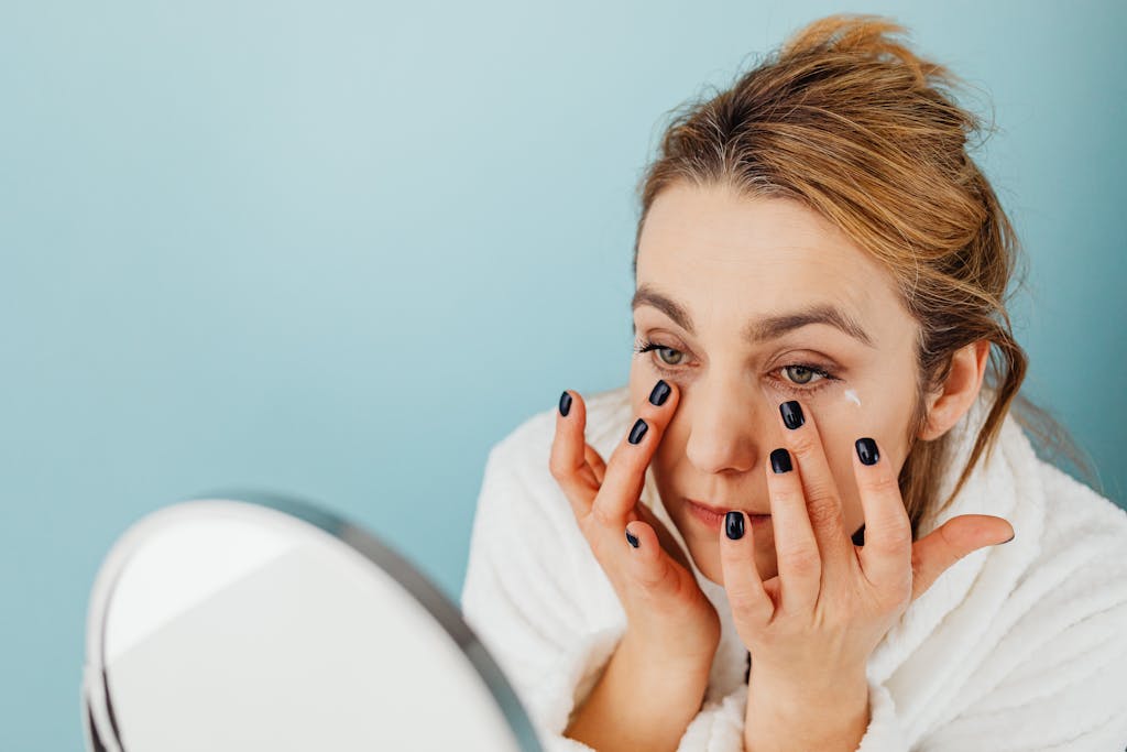 Woman applies eye cream on her under-eye area with black nails, wearing a white robe.