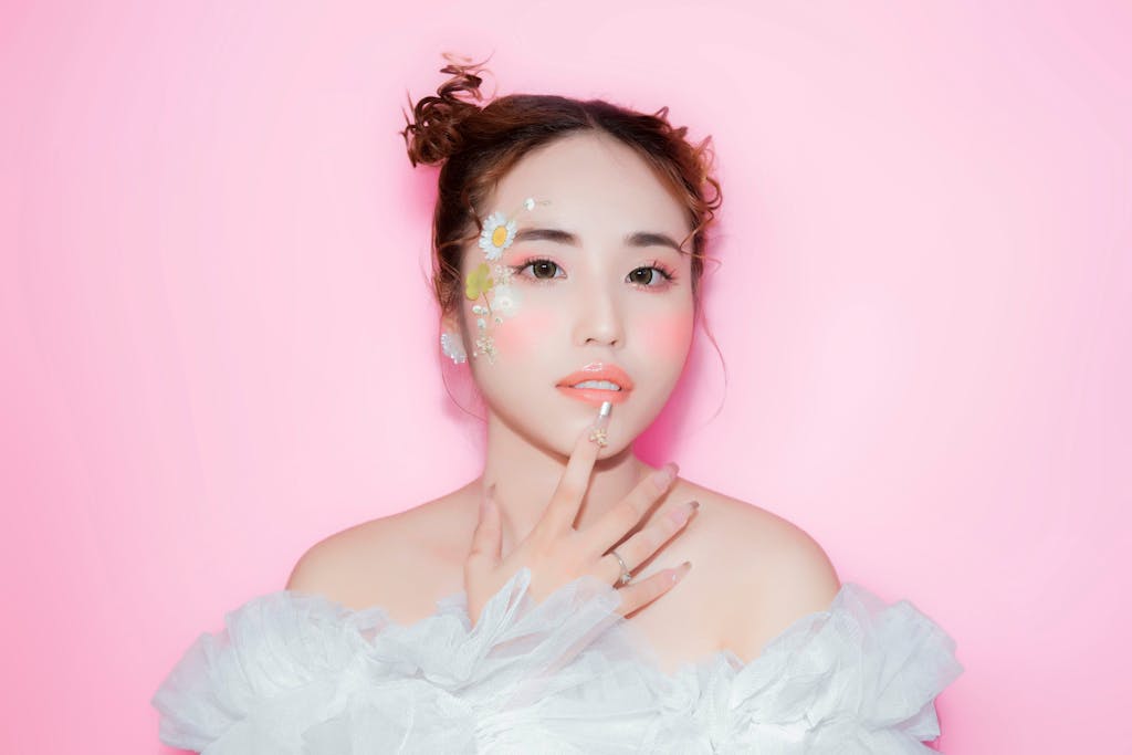Young adult woman with floral face art and chiffon dress against a pink background.