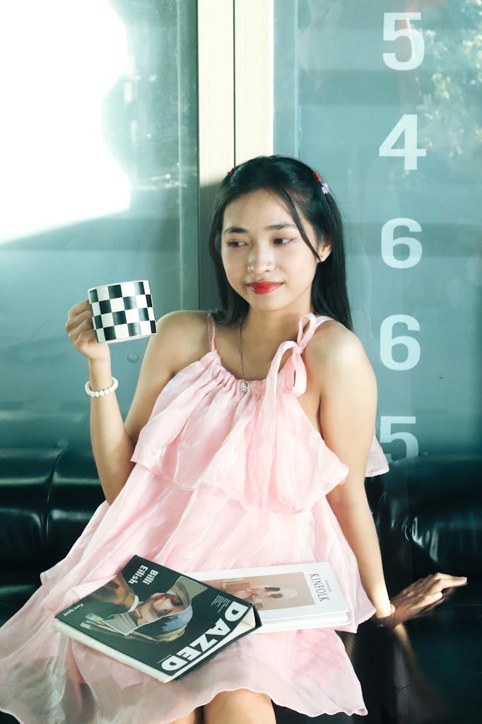Young woman in a pink dress sitting indoors with a magazine and checkered coffee cup, exuding a relaxed vibe.