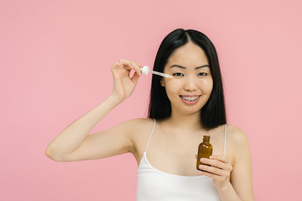 Smiling woman with braces applying facial serum on a pink background, highlighting skincare routine.