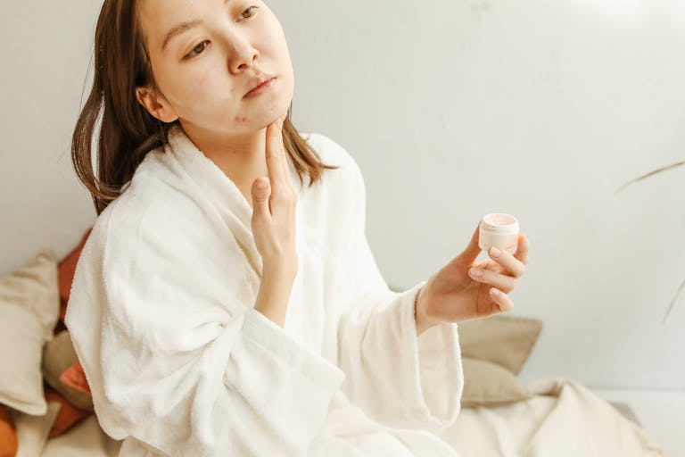An adult woman in a bathrobe applies facial cream during her skincare routine indoors.