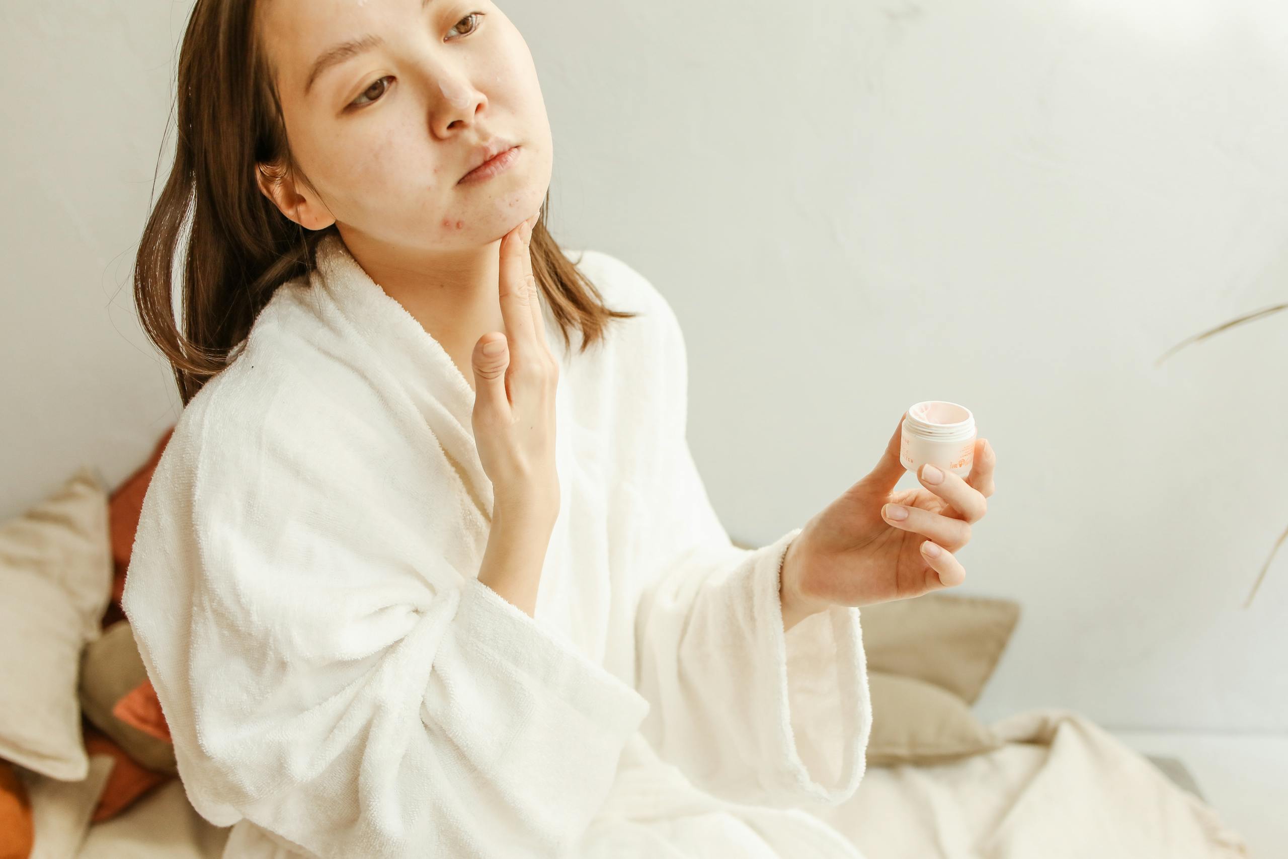 An adult woman in a bathrobe applies facial cream during her skincare routine indoors.