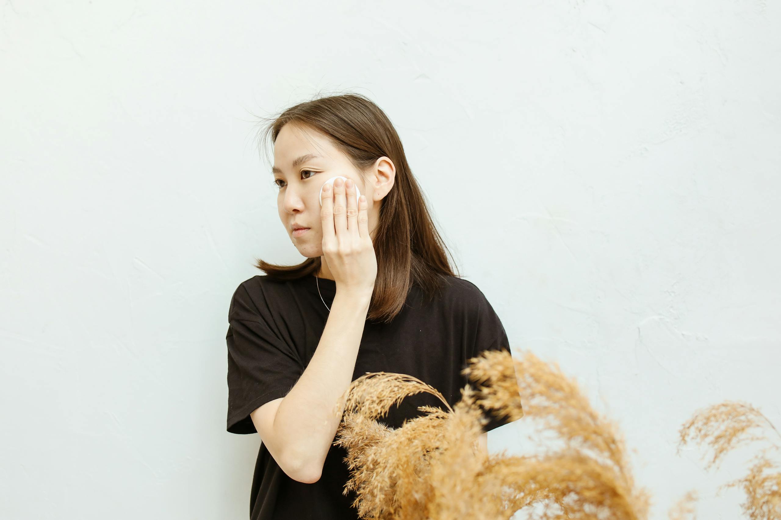 Asian woman applying skincare with a makeup remover pad, wearing a black shirt indoors.