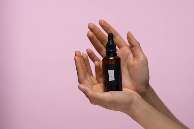 Close-up of hands holding a serum dropper bottle against a pink backdrop, symbolizing skincare.