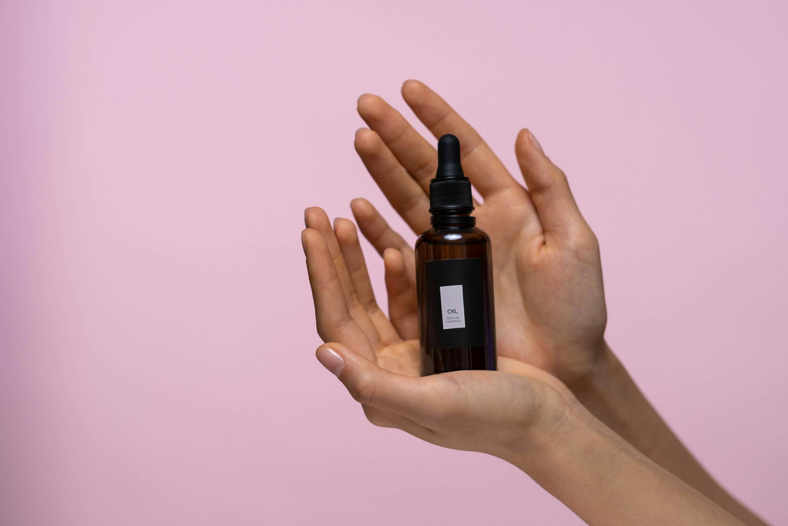 Close-up of hands holding a serum dropper bottle against a pink backdrop, symbolizing skincare.