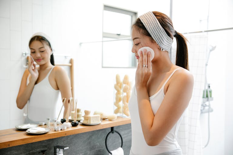 Glad Asian female wiping face with cotton pad while standing in bathroom near mirror and wooden shelf with skincare supplies during daily hygiene routine