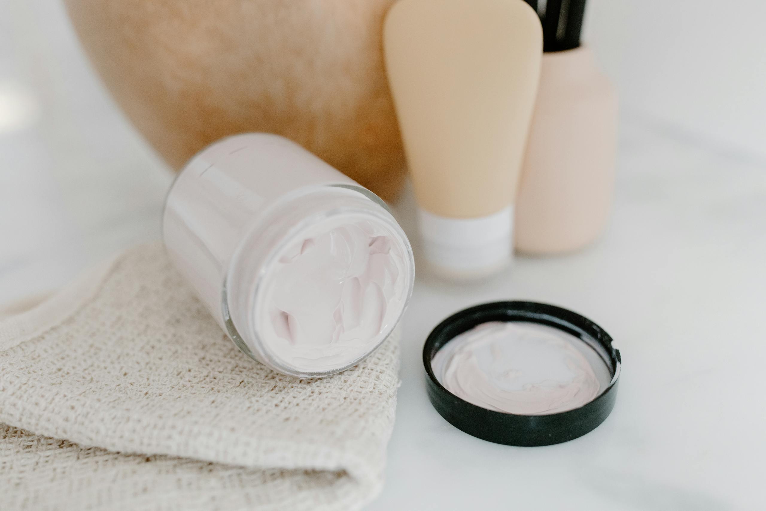 Glass jar with face cream amidst gentle skincare products on a neutral background.