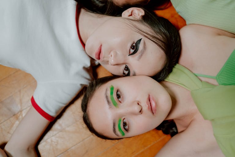 Two young women with creative neon and smoky eye makeup lying on a wooden floor together.