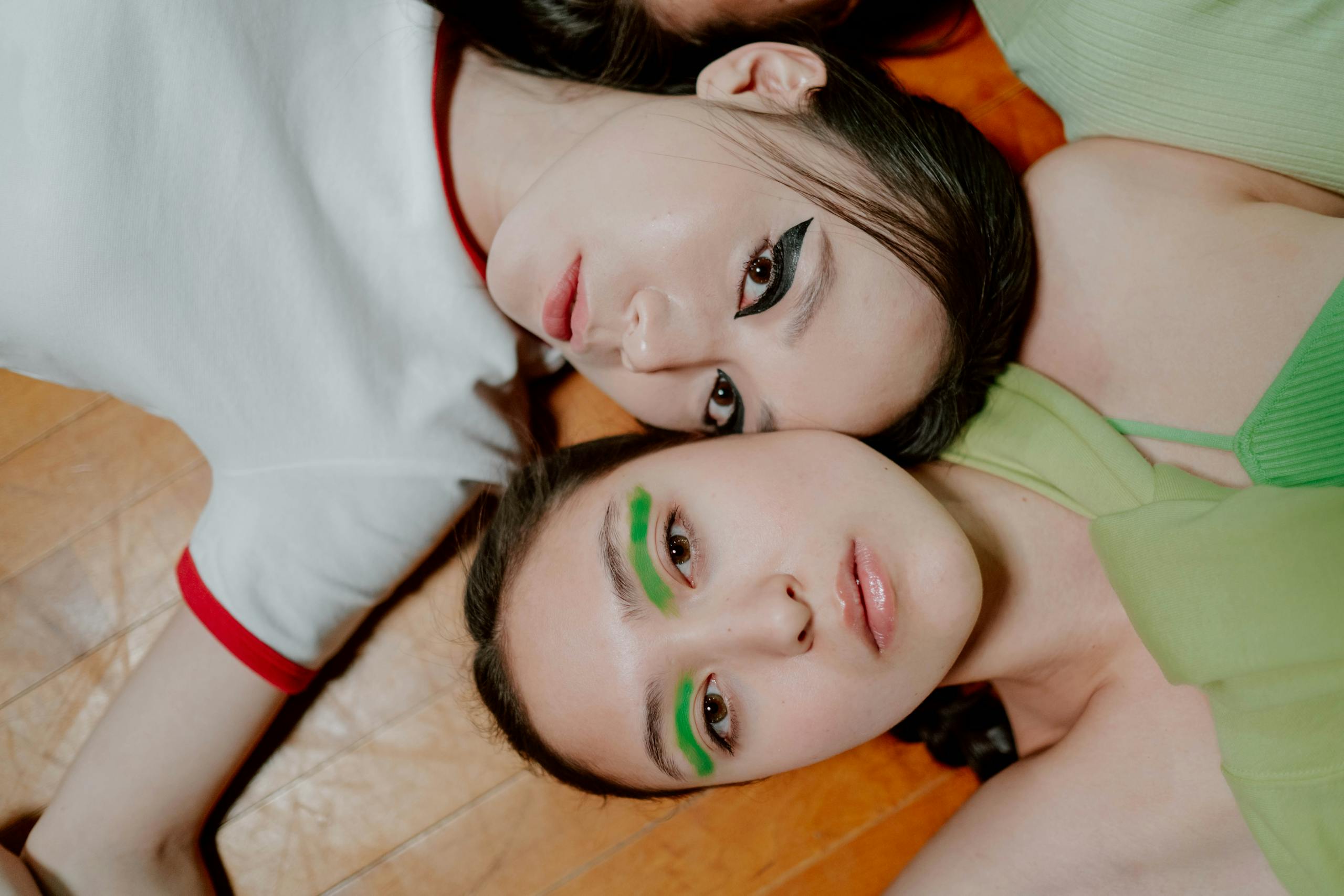 Two young women with creative neon and smoky eye makeup lying on a wooden floor together.